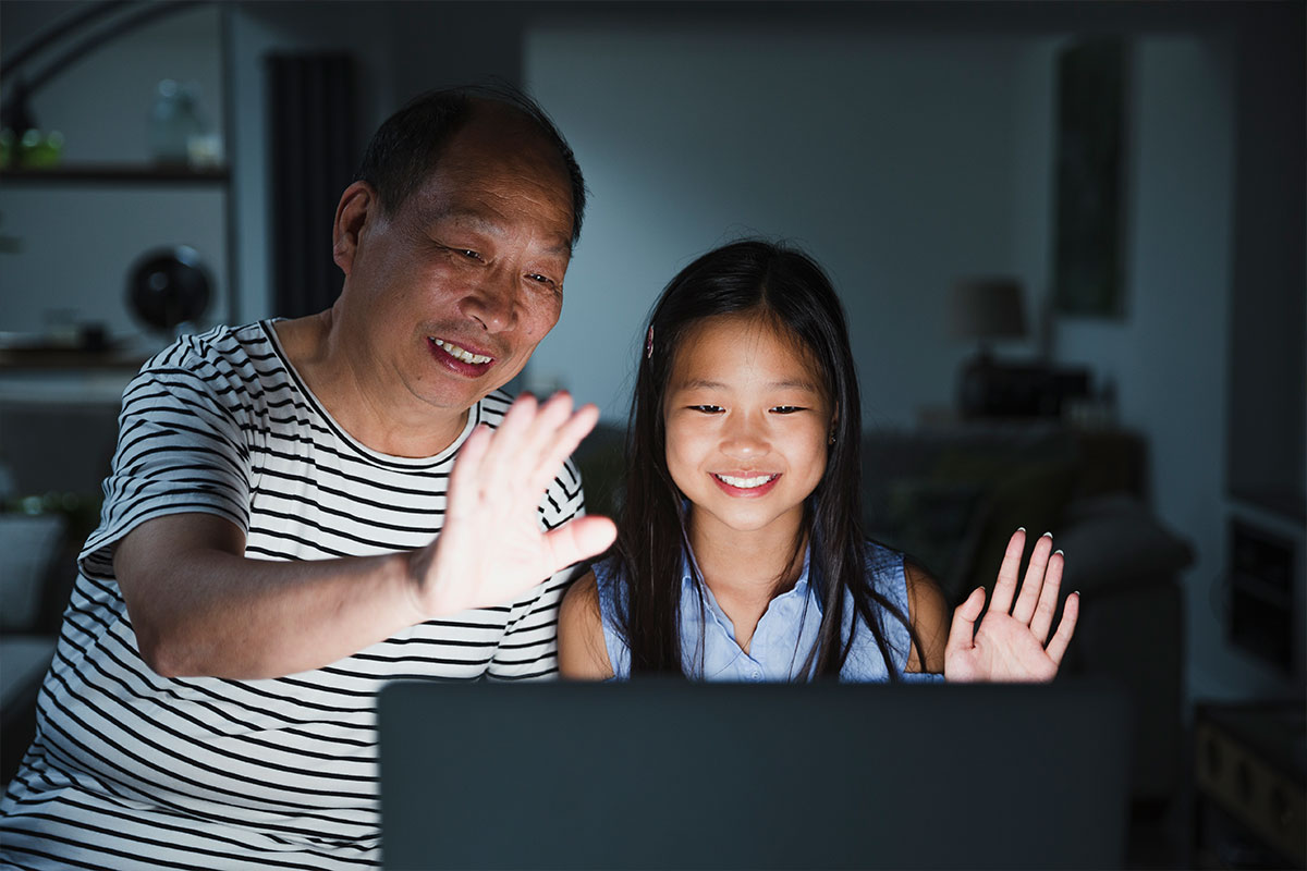 A grandpa and granddaughter smile and wave to someone on a computer screen at night