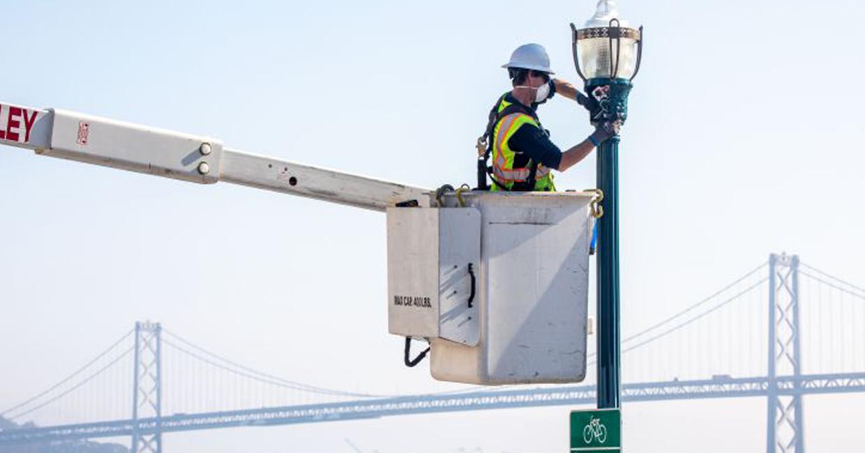 Lineworker replacing street lights with Bay Bridge in background