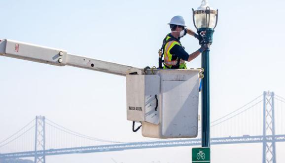 Lineworker replacing street lights with Bay Bridge in background